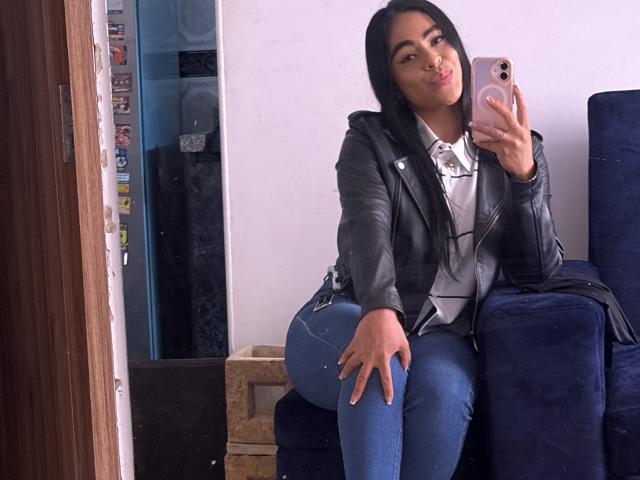 StellaBeckker with long black hair, pouty lips, black leather jacket, white shirt, blue jeans, sitting on blue chair, holding pink phone, showing curvy ass, in mirror selfie.
