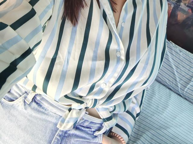 La camisa a rayas de AngelaMonticelli, con líneas verticales verdes y blancas, atada a la cintura, cubriendo sus grandes senos y pantalones vaqueros azules. Su mano descansa sobre su entrepierna..
