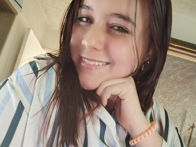 AngelaMonticelli has wet brown hair, smiling with teeth showing, wearing a striped shirt, and a peach bracelet, resting her chin on her hand, looking cute and sexy.