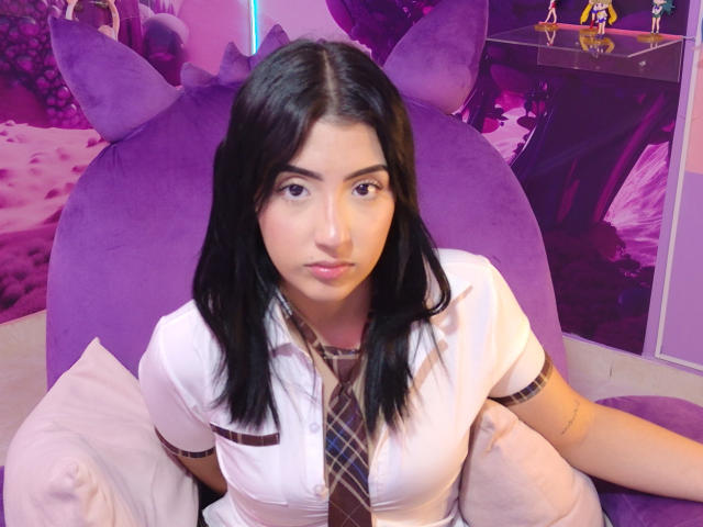 Zoey Morelli, con capelli neri, camicia bianca, cravatta a quadri marrone, seduta su una sedia viola, che guarda la fotocamera, con labbra leggermente tumefatte, piccoli tatuaggi sul braccio, sfondo viola..