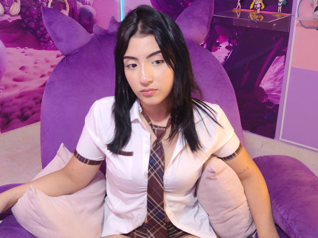 Zoey Morelli, con i capelli neri, indossa una camicia bianca, una cravatta a quadri e si siede su un divano viola, circondata da cuscini viola, guardando la fotocamera con un'espressione neutra..