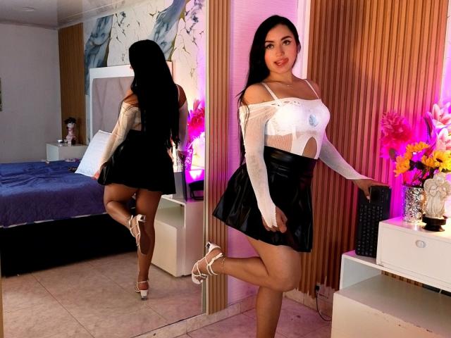 MiiaRousse with long black hair, white top showing her tits, black leather skirt, and white heels, standing in front of a mirror, holding a book, smiling. Reflection shows her ass.