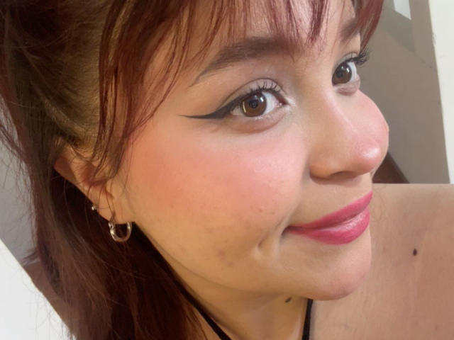 El rostro de SamyMancini, con ojos marrones delineados con delineador negro, lápiz labial rojo y una pequeña peca en su mejilla izquierda. Tiene cabello rojo, aros grandes y un top negro con tirantes..