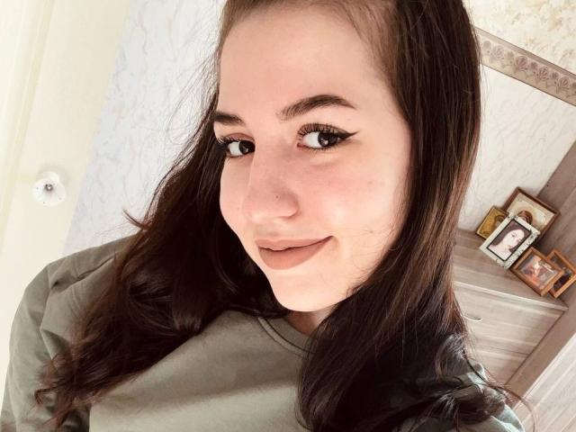 BethelVanhamme has long brown hair, brown eyes with thick eyeliner, a small nose, and a slight smile. She's wearing a green shirt, showing her cute ass.