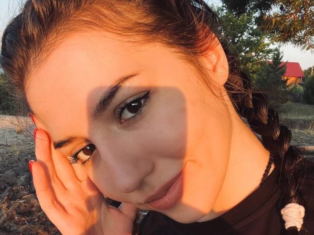 BethelVanhamme's face, sunlit, with shadow on cheek, brown eyes, braided hair, red nails, black top, slight smile, hand on face, warm light, outdoors, evening.