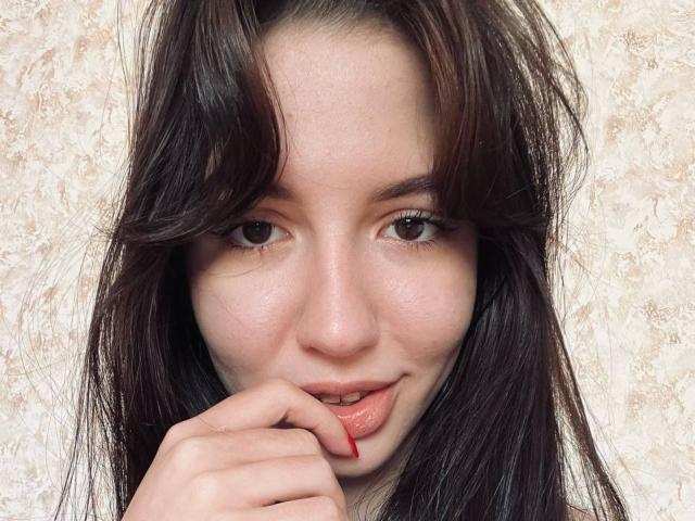 BethelVanhamme with dark brown hair, fair skin, brown eyes, slightly parted pink lips, and red fingernails touching her lower lip, showing a subtle, playful smile.