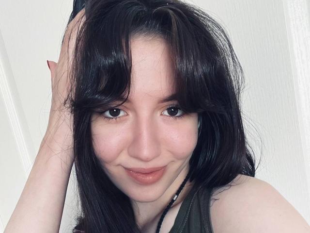 BethelVanhamme has dark, wavy hair, light skin, brown eyes, and a slight smile with pink lips. She's holding her hair up with one hand.