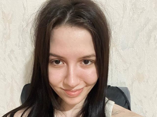 BethelVanhamme tem pele clara, cabelo preto e liso, e um sorriso discreto. Ela está olhando para a câmera com olhos castanhos, bochechas levemente rosadas e uma pequena tatuagem no braço direito..