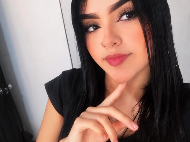 Ivy Foster hat schwarzes Haar, dunkle Augenbrauen, lange Wimpern, rosafarbene Lippen und berührt ihr Kinn mit einem Finger. Sie trägt ein schwarzes Hemd und sieht süß aus..
