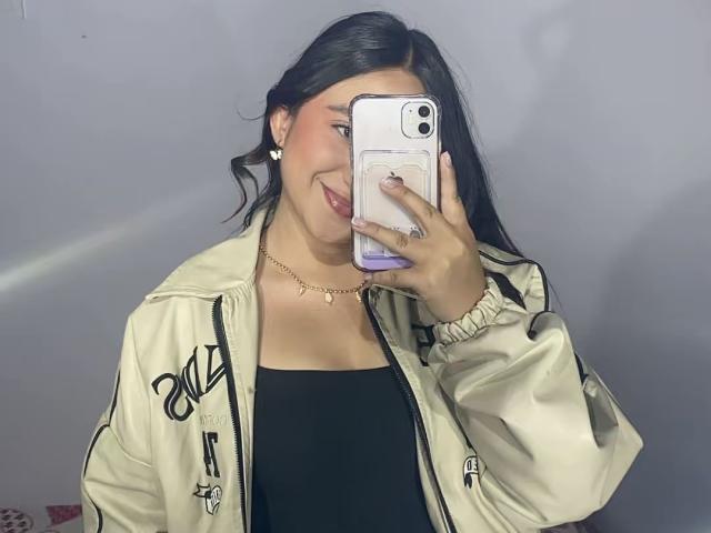 RosalieDupont with dark hair, wearing a beige jacket, black top, gold necklace, and earrings, taking a mirror selfie with a white phone, smiling subtly.
