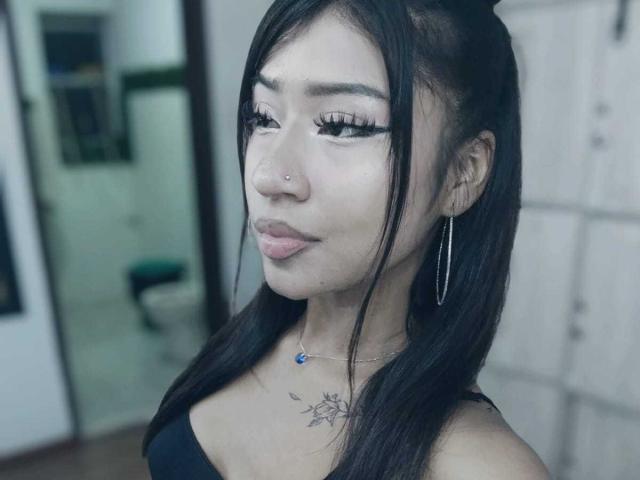 BonnieBodn, con capelli scuri, piercing al naso, orecchini a cerchio, canotta nera e collana blu, che mostra il seno e il tatuaggio sul petto. Sfondo sfocato..