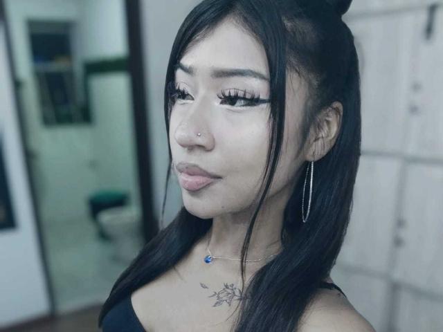 BonnieBodn a de longs cheveux noirs attachés en chignon, des cils épais, un anneau de nez, des boucles d'oreilles et un petit tatouage sous le menton, et elle porte un haut noir..
