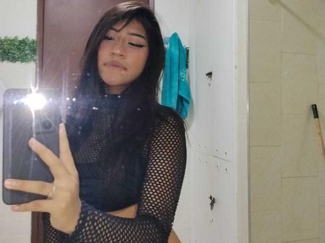 BonnieBodn, con lunghi capelli neri, indossa una maglietta nera in rete e sta scattando un selfie davanti a uno specchio del bagno. Il seno è visibile attraverso la rete. Il flash del telefono è acceso a sinistra. Sorride sardonica..