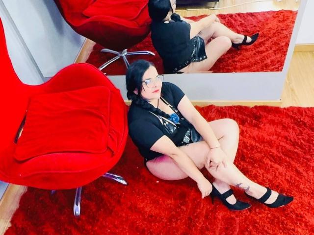 BiancaBeck, avec ses cheveux noirs, ses lunettes et son rouge à lèvres rouge, est assise sur un tapis rouge, vêtue d'une chemise noire, d'un short et de talons noirs, et son reflet est visible dans un miroir, à côté d'une chaise rouge..