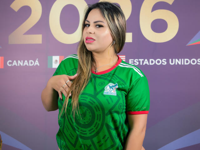 LiaAdams has long blonde hair, green soccer jersey with red trim, logo, and pattern, pink lipstick, and ring on right hand, standing in front of purple background.