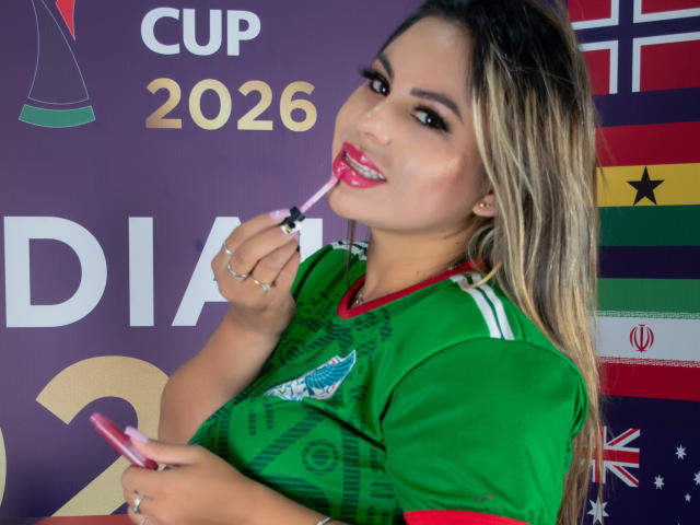 LiaAdams, avec ses cheveux blonds, son maillot de foot vert, son rouge à lèvres rouge, tenant un rouge à lèvres rose, le mordant, tenant un téléphone, souriant, exhibant sa poitrine, sexy, avec des drapeaux colorés en arrière-plan..