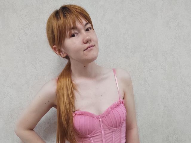 SheritaChieng, com pele clara e um longo rabo de cavalo laranja, vestindo uma blusa rosa, com babados e estilo corset, que mostra seios pequenos, e um sorriso leve e confiante, está em pé em frente a uma parede texturizada..