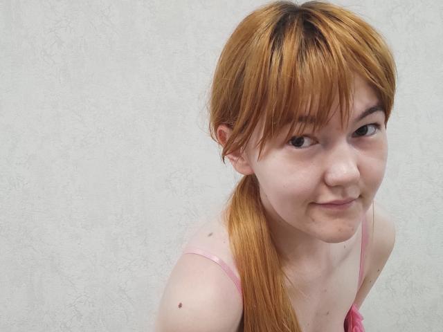 SheritaChieng, met lichtbruine vlechtjes, kleine borsten en een schattige glimlach, draagt een roze tanktop, heeft een kleine moedervlek op haar rechter schouder en een licht mollig gezicht..
