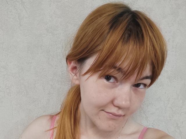 SheritaChieng met lichtbruin, steil, schouderlang haar, een roze tanktop, een kleine neus, een lichte glimlach en sproeten, die naar de camera kijkt. Haar borsten zijn niet zichtbaar..