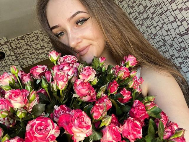 Pamela Xu, con lunghi capelli castani, tiene in mano un bouquet di rose rosa e bianche, sorridendo alla fotocamera, occhi verdi, indossa una maglietta bianca, trucco leggero, carina..