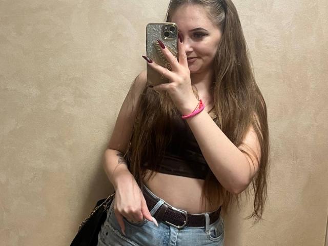 Pamela Xu, con lunghi capelli castani, indossa una canotta nera, jeans blu e un braccialetto rosa, sta scattando un selfie allo specchio, mostrando il seno, con un telefono in mano..
