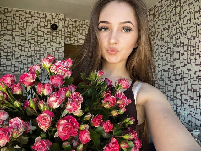 Pamela Xu, con lunghi capelli castani, labbra piene e occhi azzurri, che tiene un bouquet di rose rosa, indossa una maglietta nera con spalline bianche, in un selfie..