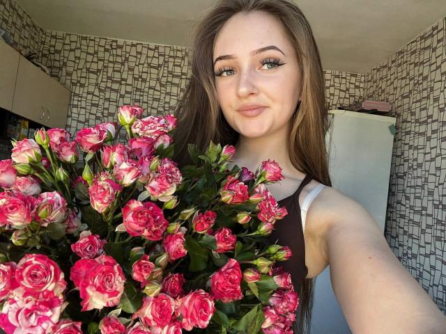 Pamela Xu, con lunghi capelli castani, occhi verdi e eyeliner allungato, tiene un bouquet di rose rosa, indossa una canotta marrone ed è sorridente in una stanza con motivi decorativi..