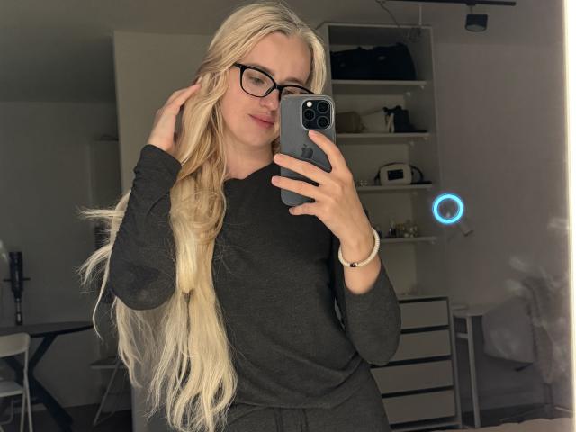 WorldClassBlondy met lang blond haar, een bril en een donkergrijs shirt met lange mouwen, die een spiegelselfie maakt, een iPhone vasthoudt, lacht en met haar rechterhand haar haar aanraakt..