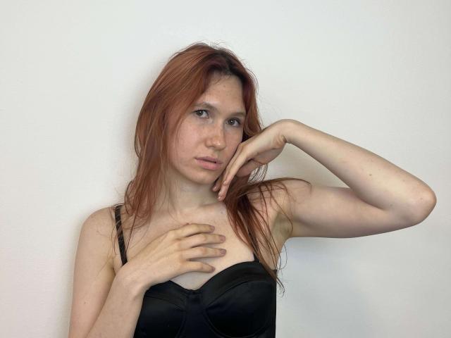 KrystaBajaj, con cabello rojo, vistiendo un sostén negro, una mano en el pecho, la otra mano tocándose la cara, piel pálida, con pecas, con una expresión ligeramente aburrida, fondo blanco, de pie..