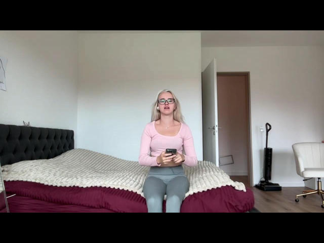 WorldClassBlondy, com cabelos loiros longos, óculos, blusa rosa e calças cinzas, sentada em uma cama, segurando um telefone, olhando para ele, com um bumbum pequeno e visível..