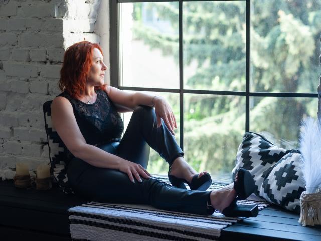 TaylorFoxT mit roten Haaren, einem schwarzen Spitzenoberteil und engen Jeans, sitzt auf einem Fensterbrett in schwarzen High Heels, die Beine gekreuzt, und lächelt dabei die Vegetation draußen an. Eine weiße Ziegelwand ist im Hintergrund zu sehen..