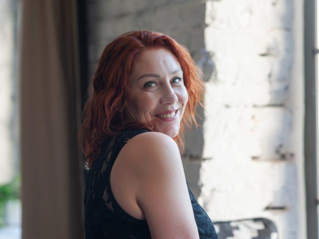 TaylorFoxT with red wavy hair, smiling, black lace sleeve, fair skin, looking over shoulder, showing her tits, standing near a white brick wall, natural light, smiling.