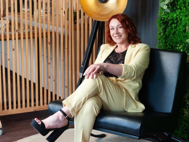 TaylorFoxT with red hair, smiling, wearing a yellow suit, black lace top, and black heels, sitting on a black chair, crossed legs, wood and greenery background.