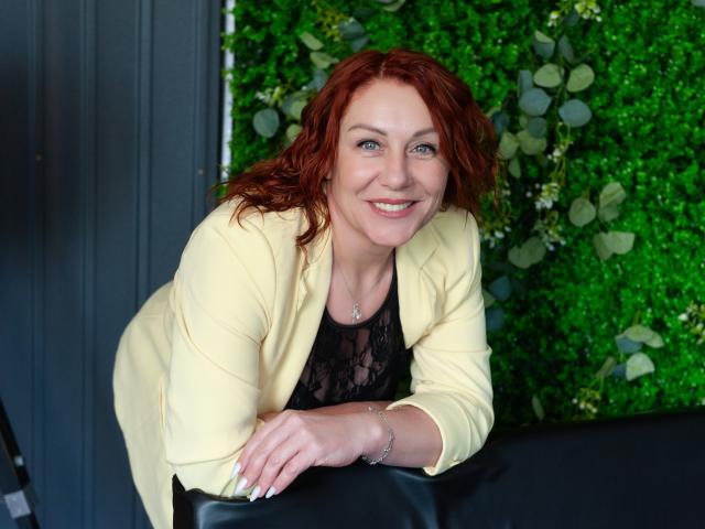TaylorFoxT with red hair, smiling, wearing a yellow blazer over a black lace top, leaning on a black chair, green wall background, blue eyes, and a silver necklace.