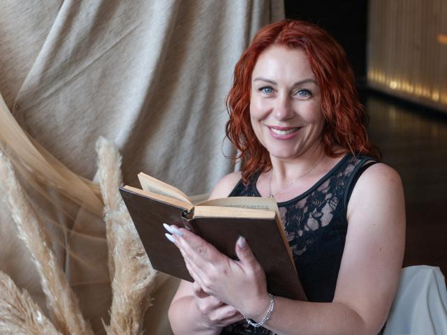 TaylorFoxT with red hair, black lace sleeveless top, holding open book, smiling, white nails, silver bracelet, standing near pampas grass, indoor setting.