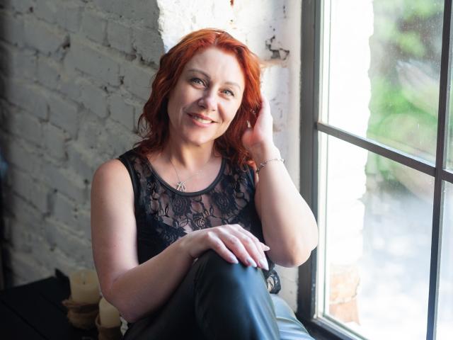 TaylorFoxT, rothaarige Frau, lächelnd, Oberteil aus Spitze, schwarze Lederhose, sitzend am Fenster, Hand auf dem Kopf, weiße Ziegelwand, weiches Licht, Grün draußen, süß, sexy, attraktiv..