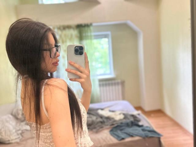 Merrie Putnam, con cabello negro largo, gafas y una blusa blanca de encaje, se toma una selfie frente al espejo, mostrando su espalda y trasero. Se ven una cama y ropa en el fondo..