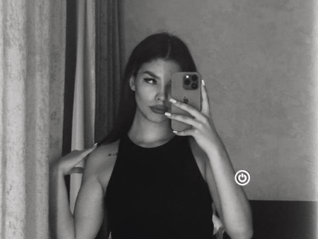Selfie in bianco e nero di MerriePutnam con lunghi capelli scuri, che indossa una canotta nera senza maniche, tiene un telefono nella mano destra, guarda la fotocamera, mostrando il suo sedere..