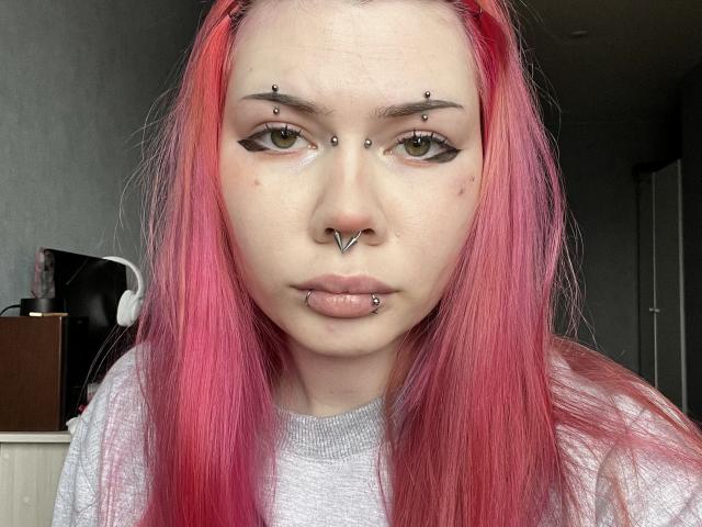 ToriHananocha tiene el cabello rosa, ojos verdes y múltiples perforaciones faciales: en la nariz, el labio y sobre las cejas. Lleva una camiseta gris y tiene una expresión seria..