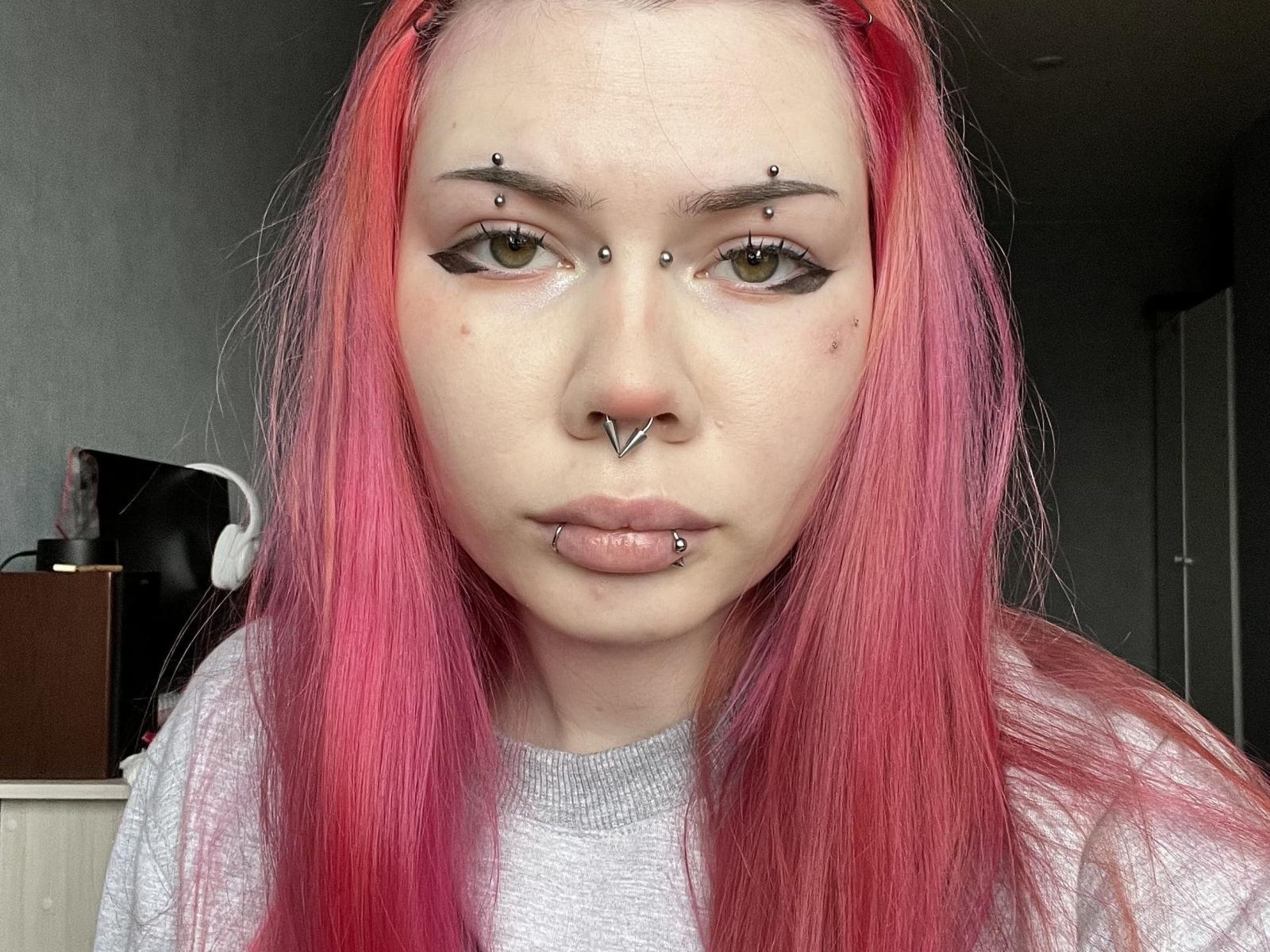 ToriHananocha tiene el cabello rosa, ojos verdes y múltiples perforaciones faciales: en la nariz, el labio y sobre las cejas. Lleva una camiseta gris y tiene una expresión seria..
