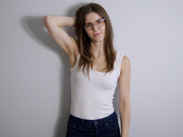 GlamMegi com óculos, camiseta branca e calça jeans escura, braço levantado, cabelo castanho, sorrindo, em pé em frente a uma parede branca lisa, com uma sombra discreta à esquerda..