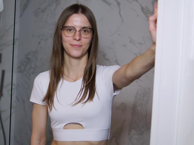 GlamMegi, con cabello castaño lacio, gafas y un top blanco corto con un corte, está de pie en un marco de puerta, sonriendo ligeramente. Tiene pechos pequeños y una figura delgada..