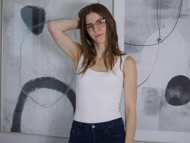 GlamMegi con gafas, cabello castaño, camiseta blanca sin mangas y jeans oscuros, brazo levantado, mirando a la cámara, sonrisa sutil, busto de tamaño medio, complexión delgada, pose casual..