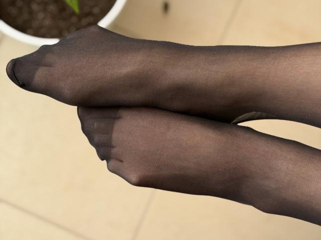 Sanella's black sheer pantyhose-covered feet are crossed at the ankles, showing her toes and slightly curved toes with a small black dot near the tip of her right toe.