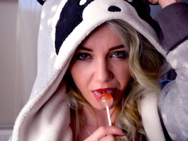 VeraNoires with blonde hair, blue eyes, and red lipstick, wearing a black and white cow hoodie, sucking on an orange lollipop, her hand in the hood.