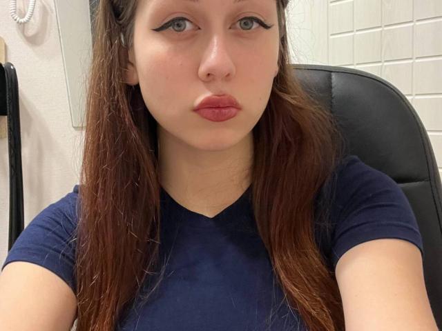 Carrie Sorenson, mit heller Haut und braunen Haaren, trägt ein dunkelblaues Hemd, hat volle, rosafarbene Lippen und schwarzen Eyeliner und sitzt auf einem schwarzen Stuhl..