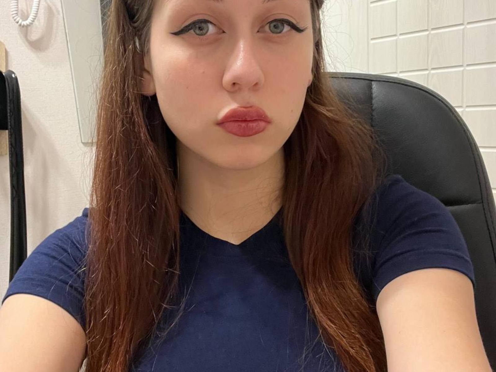 Carrie Sorenson, mit heller Haut und braunen Haaren, trägt ein dunkelblaues Hemd, hat volle, rosafarbene Lippen und schwarzen Eyeliner und sitzt auf einem schwarzen Stuhl..