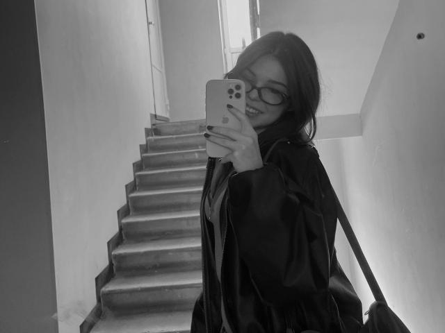 Autoportrait en noir et blanc d'AlexXas portant des lunettes, souriant, tenant un téléphone, portant une veste sombre et un sac à dos, debout sur des marches, montrant le haut du corps, avec une coque de téléphone visible..
