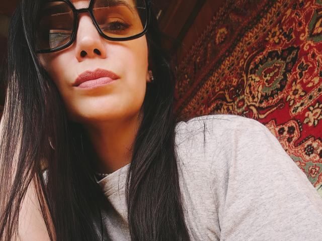 AlisaLowq mit langem, schwarzem Haar, die dicke, schwarze Brille trägt und ein graues T-Shirt anhat, hat rosafarbenen Lippenstift und ein leichtes Lächeln und blickt nach unten in die Kamera..