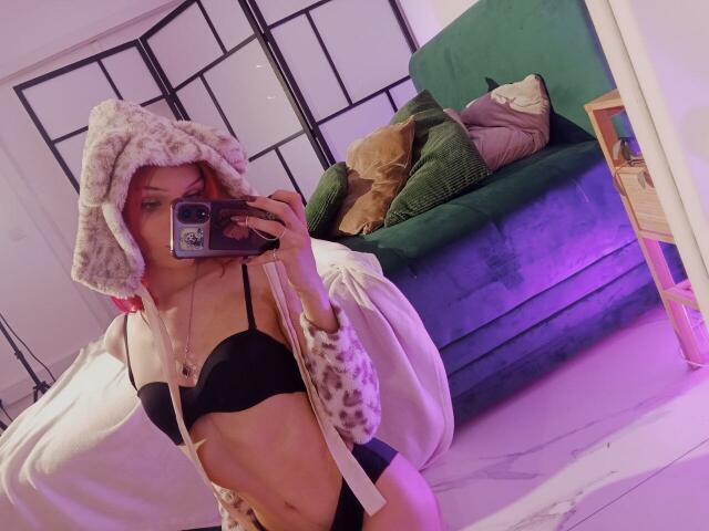 KiranaHayes in a mirror selfie, wearing a fluffy white hood, black bra, and leopard print sleeves, with her phone covering her face, exposing her tits and flat stomach.